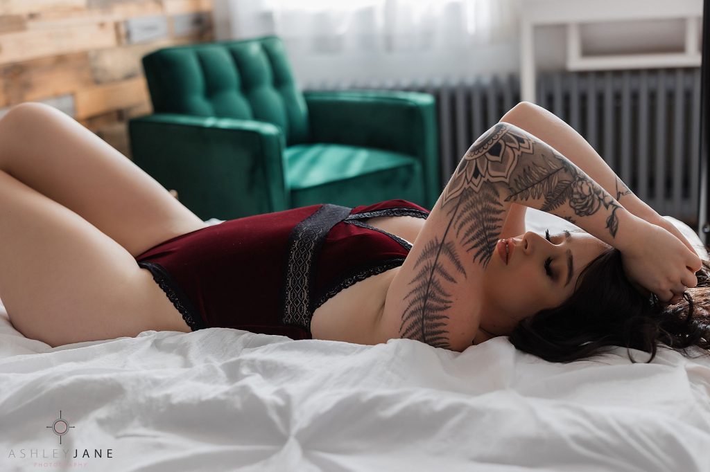 Boudoir model wearing dark red lingerie with black lace trim laying on a bed with her hands in her hair.