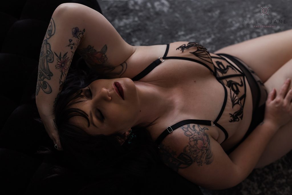 Boudoir model wearing black lacy lingerie with a dragon design. Sitting on a grey mottled carpet leaning back against a black couch, hand in her hair.
