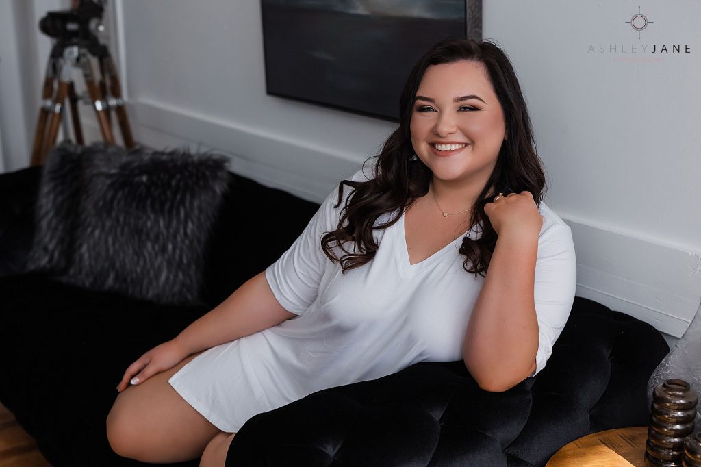 A boudoir model in a long white t-shirt sitting on a black couch, smiling at the camera.
